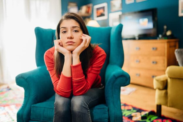 A woman sitting on a blue chair in a living room, reflecting teenage anxiety.